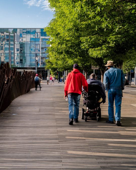 A person using a motorized wheelchair is flanked by two pedestrians on an urban sidewalk clad in wooden boards and shaded by leafy trees