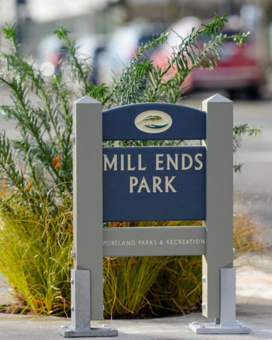 A wooden sign reading \"Mill Ends Park Portland Parks & Recreation\" stands on a concrete ring enclosing a small planter filled with an evergreen bush and light green grass