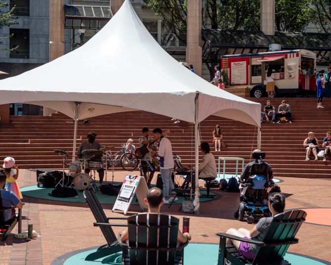 white tent with band surrounded by people in chairs, people sitting on steps in an urban courtyard