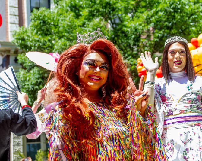 two drag queens smile and wave at the camera in front of yellow and red balloons