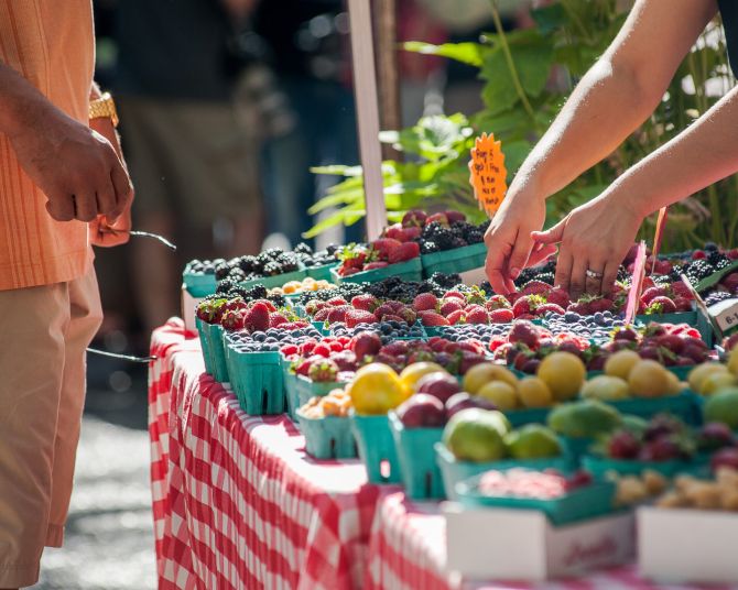 two people, a vendor and a shopper, stand over a table full of berries and local fruits