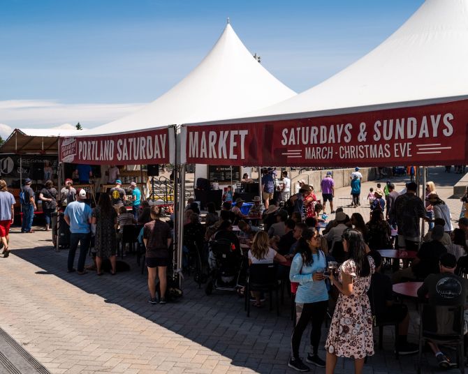 People stand under a canopy with signs reading "Portland Saturday Market Saturdays & Sundays March - Christmas Eve"