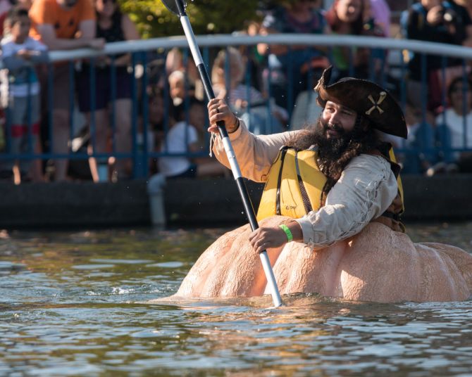 a person paddles a boat made out of a giant pumpkin with spectators watching on the shore