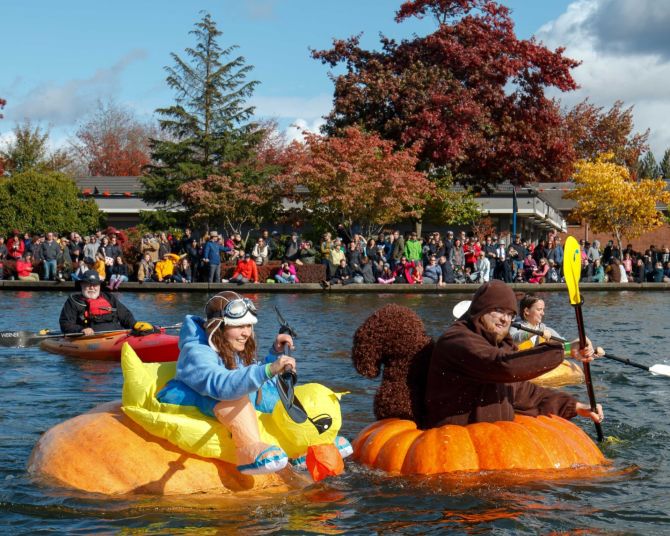 people in costumes paddle boats made of giant pumpkins on a lake while spectators watch on the shore