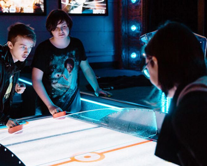three youths play a game of air hockey with brightly backlit posters in the background