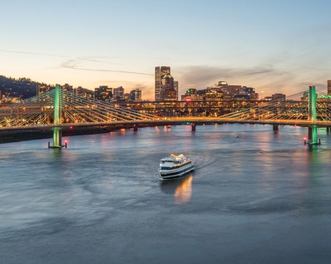 a Portland Spirit cruise boat on the Willamette River at dusk with the bright lights of downtown Portland in the background