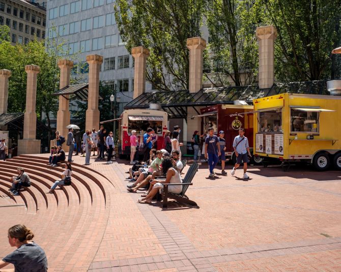 people sit in chairs and on steps in front of food carts in a brick courtyard in the center of an urban downtown area