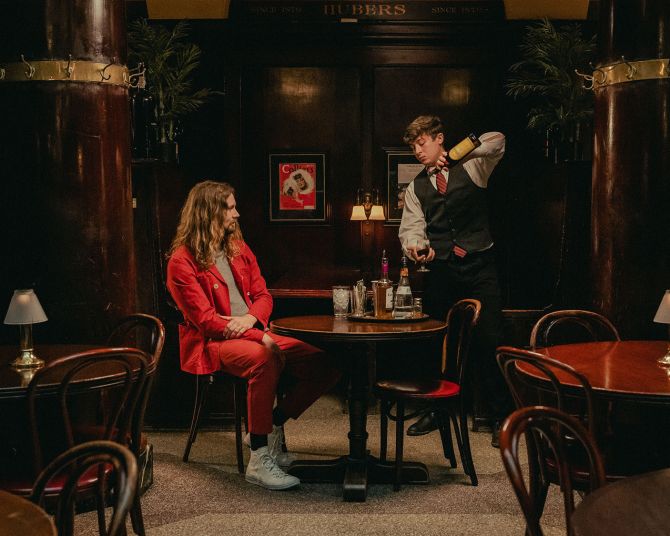 a server in an atmospheric restaurant prepares a cocktail for a man in a red suit seated at a table