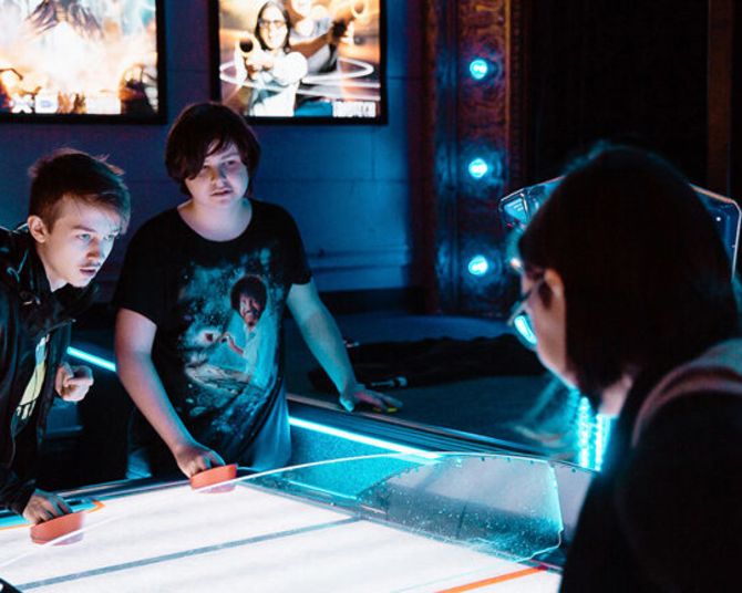 three youths play a game of air hockey with brightly backlit posters in the background