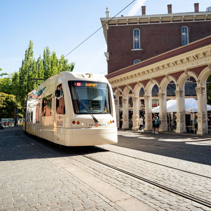 a light rail train on a cobblestone road passes the historic Saturday Market entry arches