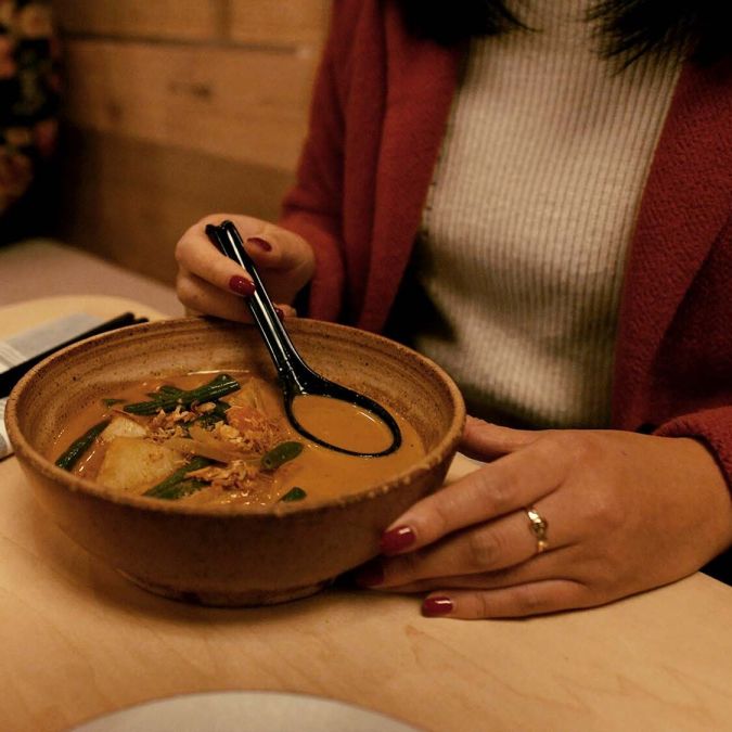 A person sitting at a table holds a ramen spoon in a bowl