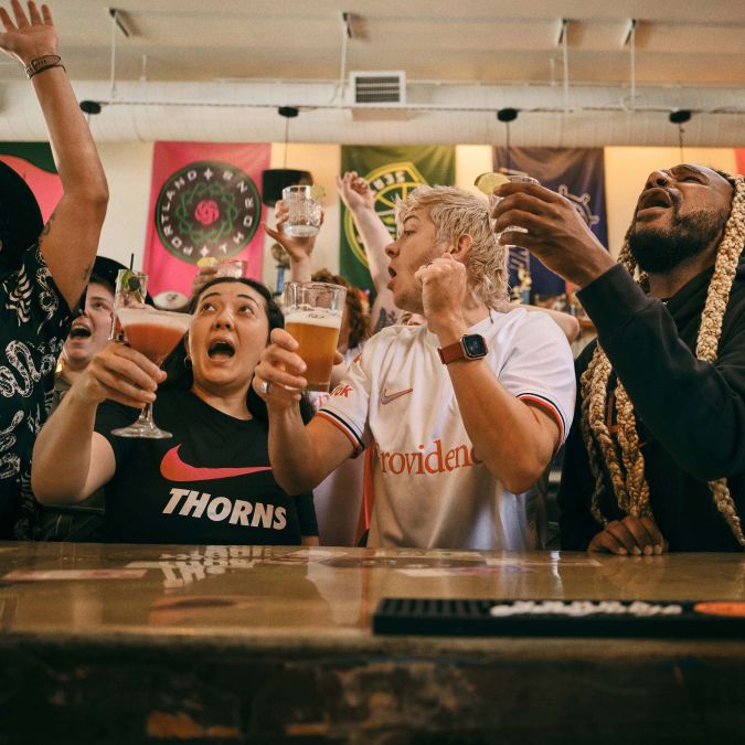 a crowd of people cheer while raising glasses in front of a bar while watching a game