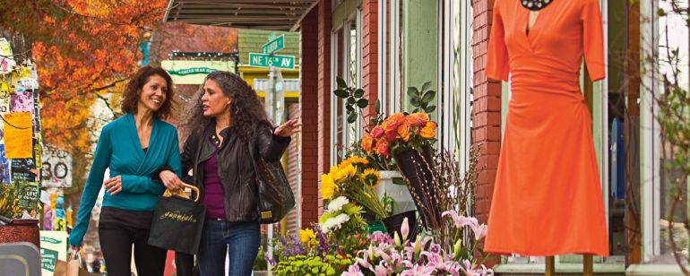 Two shoppers walking down the sidewalk in front of shops displaying flowers and clothing on Northeast Alberta Street