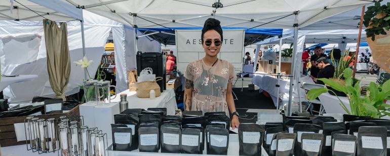 a woman smiles behind a display of beauty products in an outdoor market