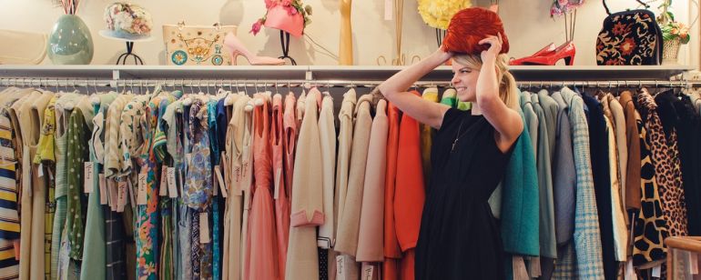 a woman trying on a hat in front of racks of vintage clothing