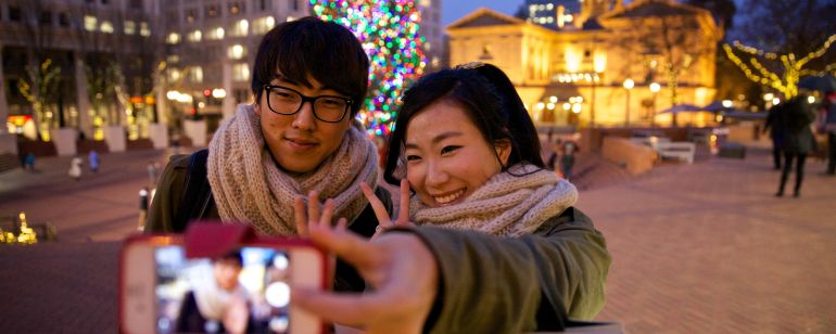 A couple poses for a selfie in front of a Christmas tree and a historic building