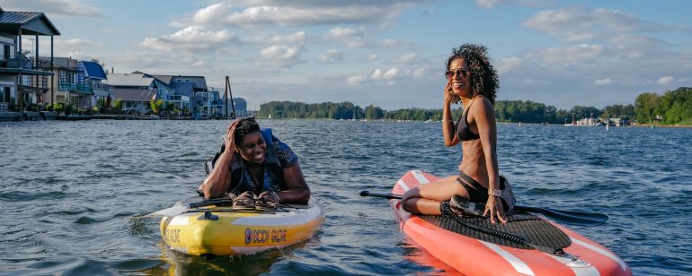 Two women on paddleboards in a river with houseboats in the distance