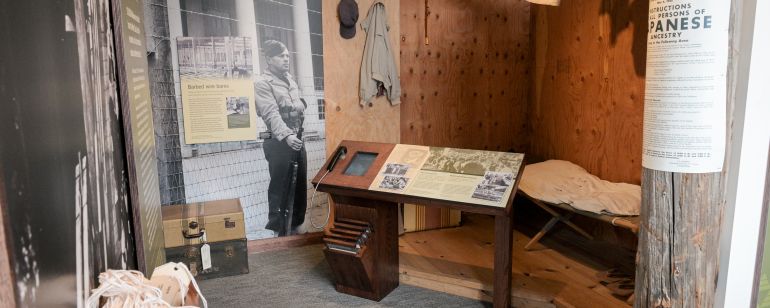 small living space including a cot and hung up laundry with image of a barbed wire fence surrounding, depicting Japanese internment living quarters