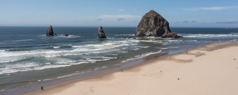 the Oregon coast on a sunny day, with waves rolling toward a sandy beach, around huge rock formations, beneath a blue sky