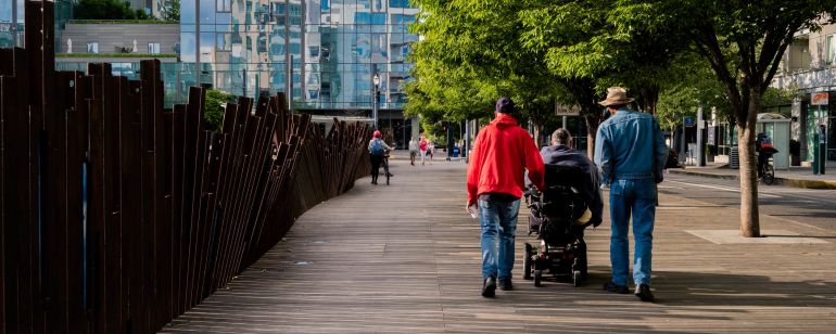 A person using a motorized wheelchair is flanked by two pedestrians on an urban sidewalk clad in wooden boards and shaded by leafy trees
