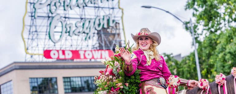 woman in hat and chaps ride a rose decorated horse in a parade with Portland\'s white stag sign in the background