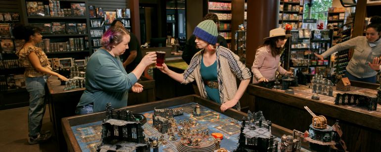 Two friends clinking glasses over a large table-top board game.