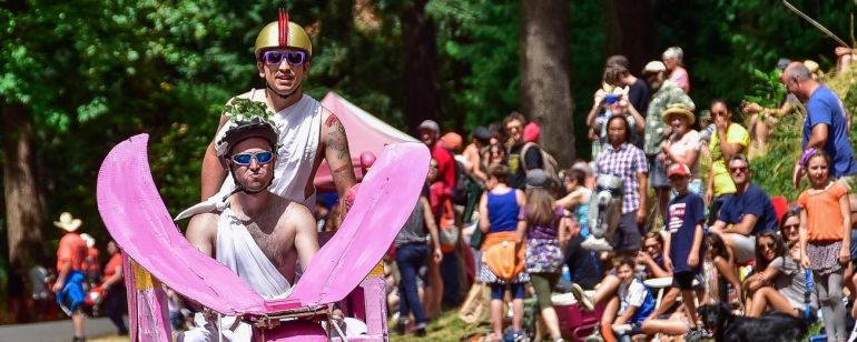 two people wearing sunglasses and togas ride a pink elephant-shaped soapbox derby cart