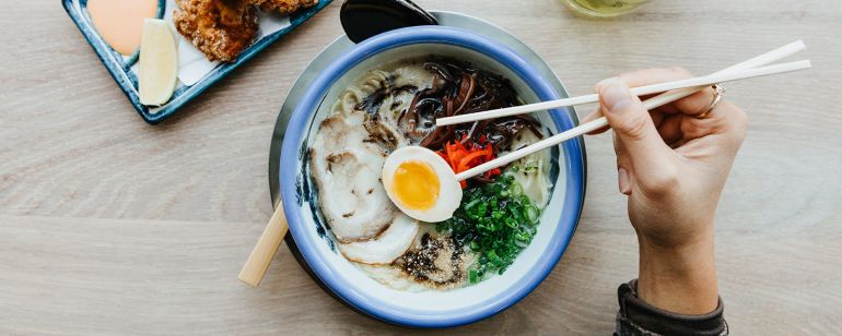 a hand holding chopsticks hovers over a bowl of ramen. a mug of green tea and a plate of fried chicken are next to the ramen bowl on a table.