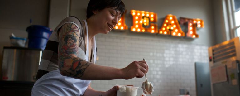 A chef tops a strawberry dessert with whipped cream in front of a neon sign reading \"MEAT\"