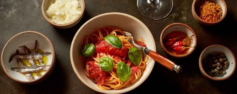an overhead view of a bowl of pasta, garnishes in ramekins and a glass of wine