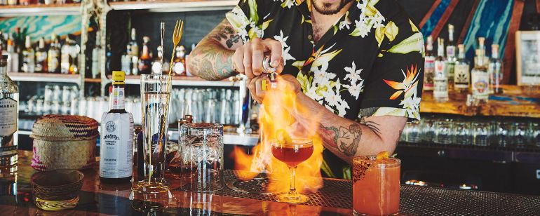 a tattooed bartender in a Hawaiian shirt sets a cocktail on fire