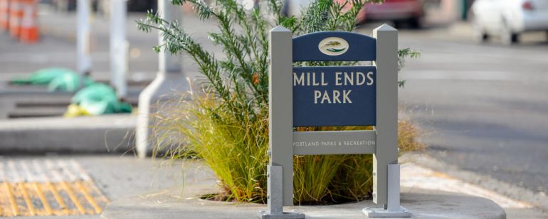A wooden sign reading \"Mill Ends Park Portland Parks & Recreation\" stands on a concrete ring enclosing a small planter filled with an evergreen bush and light green grass