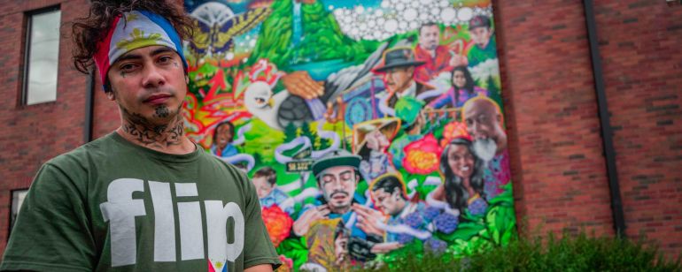Talilo Marfil, wearing the flag of the Philippines as bandana and a shirt with the word, \'flip,\' (a slang term for Filipino) stands in front of a colorful mural depicting his likeness.