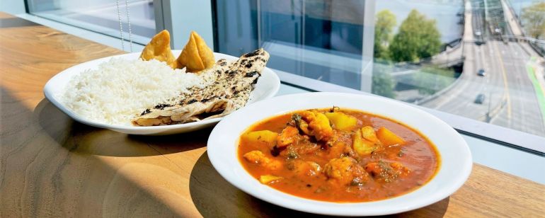 A white plate of aloo gobi sits next to a platter of rice, samosas and naan on a wooden table in front of a large window.