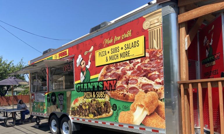 a red and green food cart with a sign advertising pizza and subs beneath clear blue skies