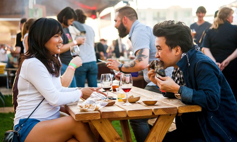 two people eating food and drinking wine at a picnic table during a busy food festival