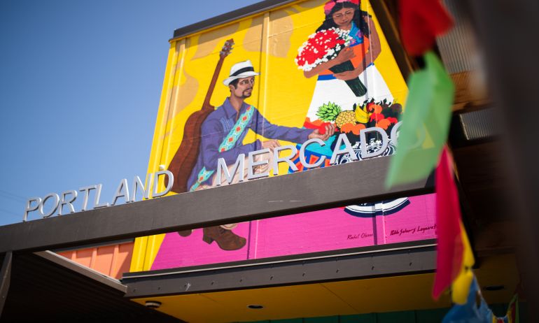 A sign reading \"Portland Mercado\" in front of a brightly painted mural showing a Latin American dancer and musician