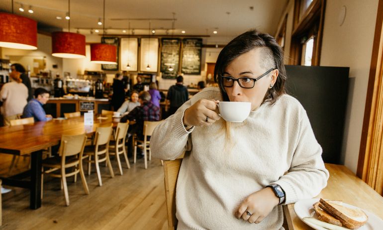 a white person with long hair on one side and a shaved head on the other side, sips from a coffee mug in a warmly lit bakery.