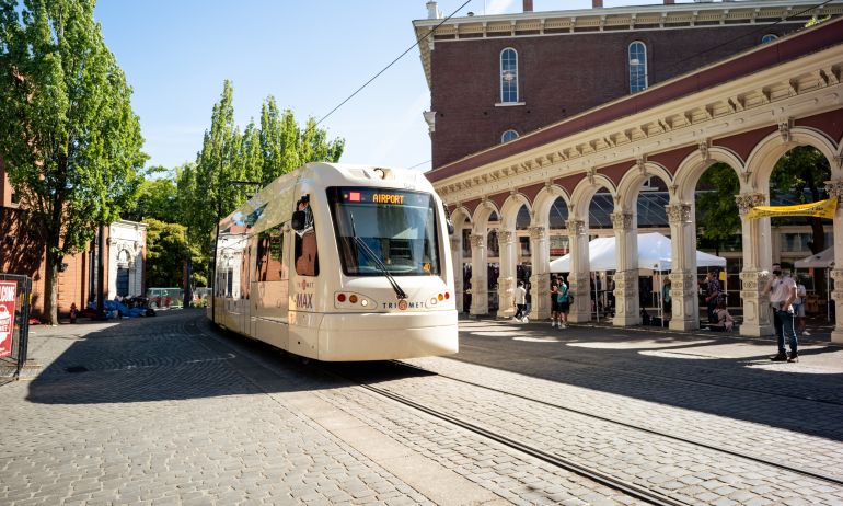 a light rail train on a cobblestone road passes the historic Saturday Market entry arches