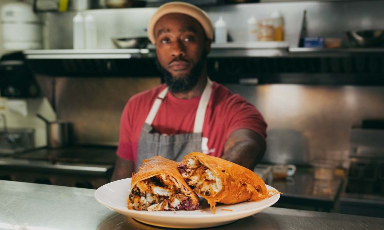 a Black man in a red shirt, yellow beanie and apron presents an overstuffed burrito on a plate