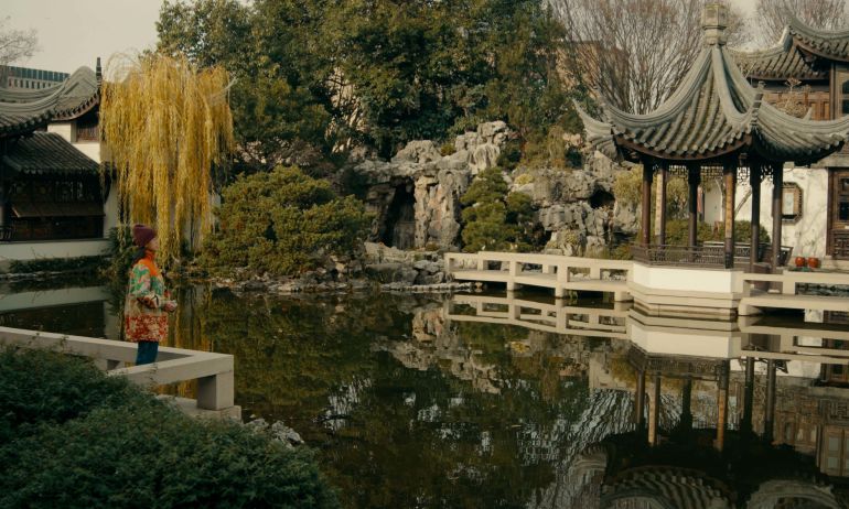 a woman holds a camera at a traditional Chinese garden with a pond, pavilion and trees