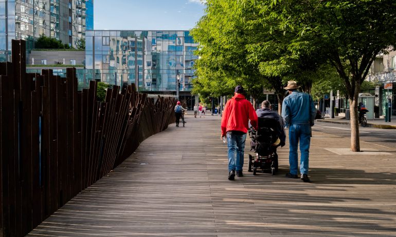 three people — one using a motorized wheelchair, two on foot — traverse a wide wooden walkway beneath flourishing green trees
