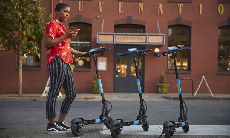 person with phone in hand stands with three electric scooters in front of a storefront