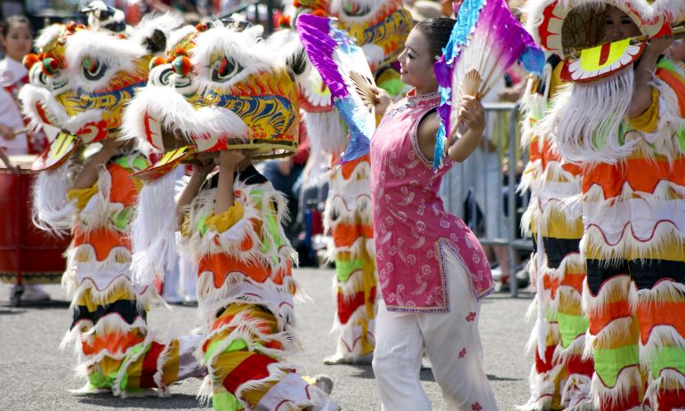 women dances with large fans outside on a city street surrounded by people in colorful dragon costumes