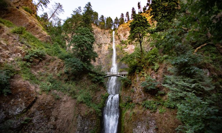 sunset hits the top of the mountain where a large waterfall bursts through the trees and cascades down the hillside, with a small bridge overlook
