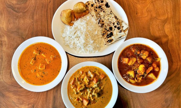 Three dishes of curries and aloo gobi sit next to a platter of rice, samosas and naan on a wood table.