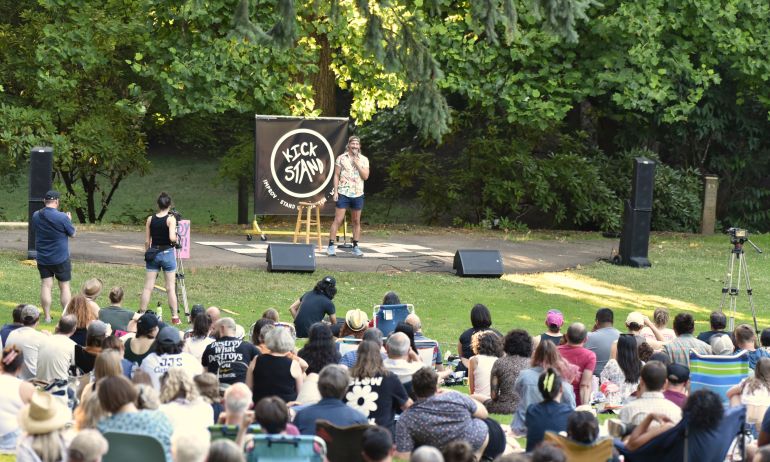 Blond comedian on stage performing in front of large crowd sitting in a park