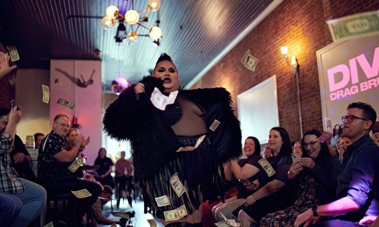 A woman in a sheer bedazzled body suit and black feather jacket performs amidst a crowd showering her with dollar bills.