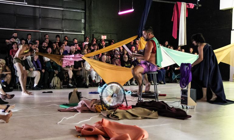 a person sits on a stool in front of an audience with colorful clothing strewn about the stage