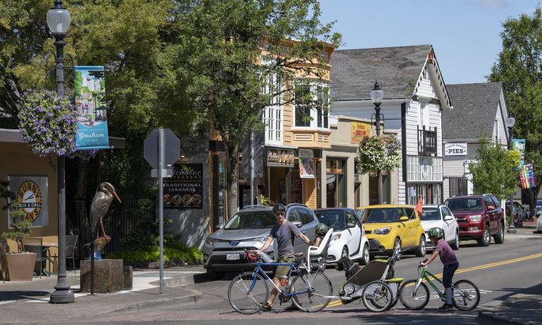 a man walking a bike across the street with a trailer with a dog attached, followed by a child on a bike, at a crosswalk with cars waiting